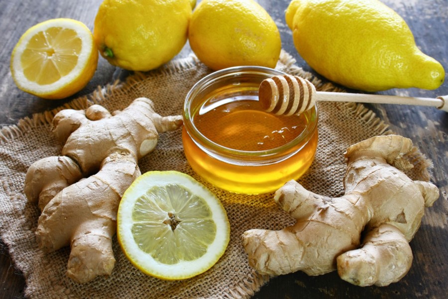 Na zdjęciu widać świeży imbir, cytryny oraz słoik miodu z drewnianą pałeczką do nabierania. Składniki te leżą na jutowej tkaninie, na drewnianej powierzchni. Całość tworzy naturalną kompozycję, często kojarzoną z domowymi sposobami na zdrowe gardło.